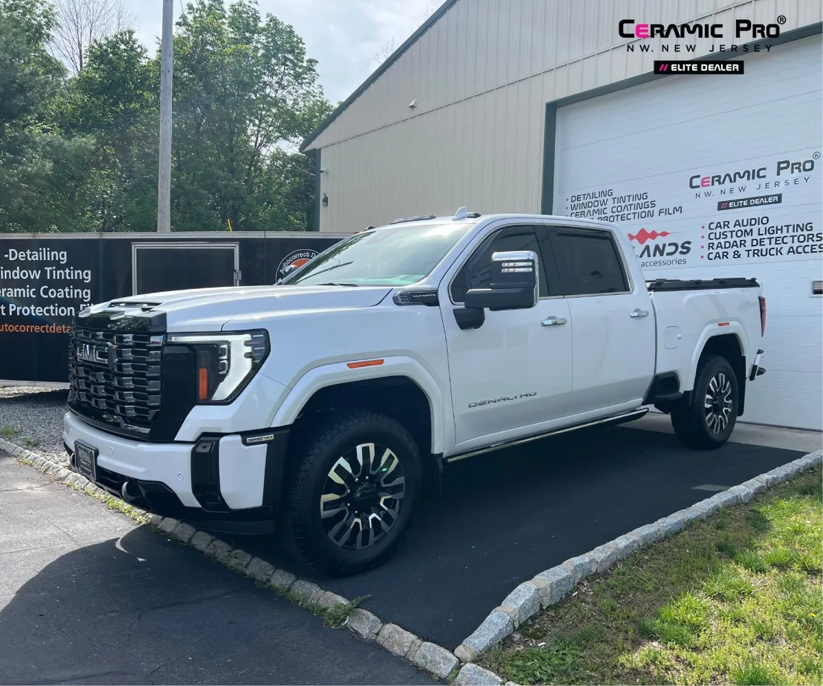 Front three-quarter view of a white GMC Sierra Denali HD truck parked outside a Ceramic Pro shop garage door.